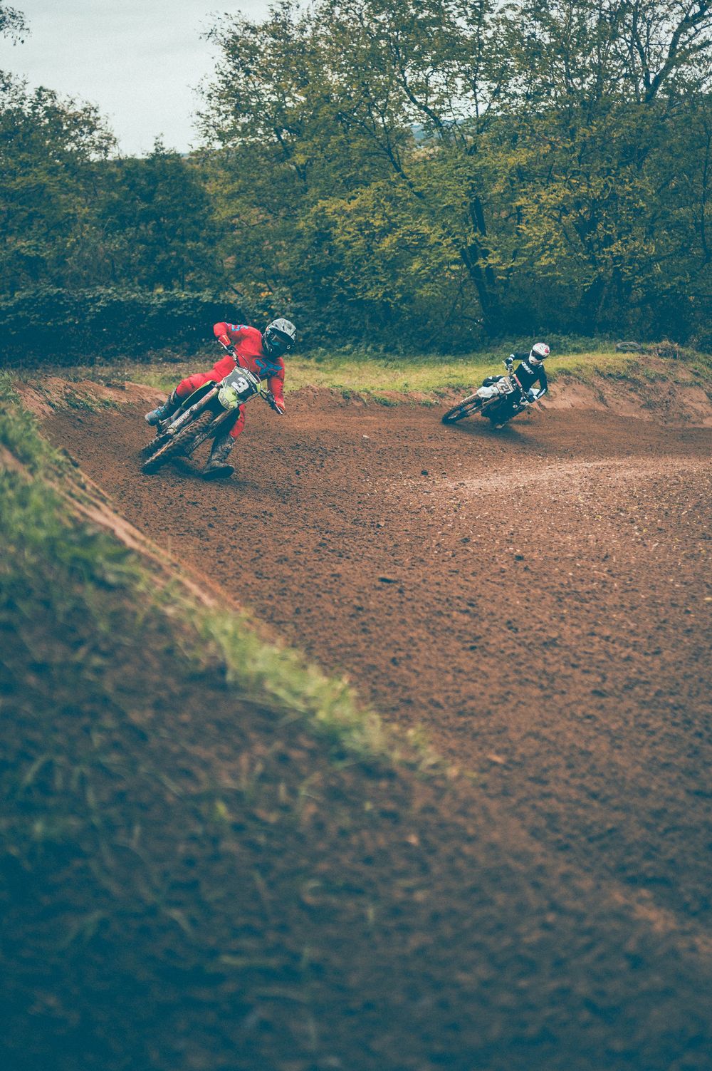 Two motocross riders in red and dark gear lean into a banked dirt turn on a muddy off-road track surrounded by green trees.