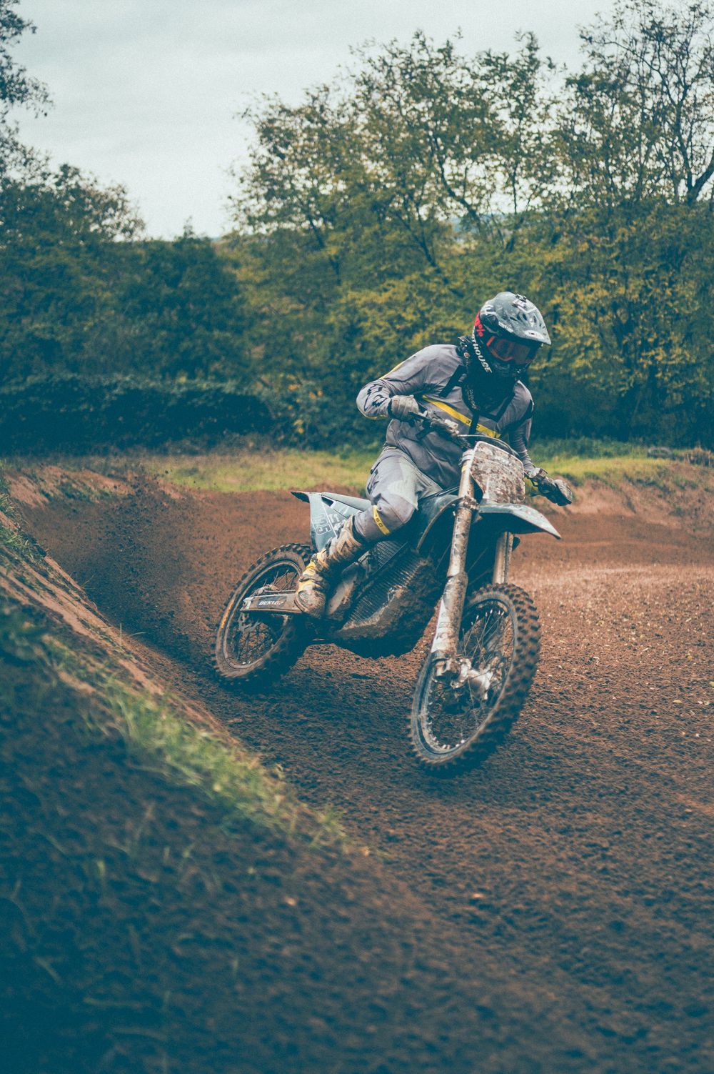 Motocross rider in a helmet and riding gear leans a dirt bike through a banked turn on a muddy off-road track surrounded by trees.