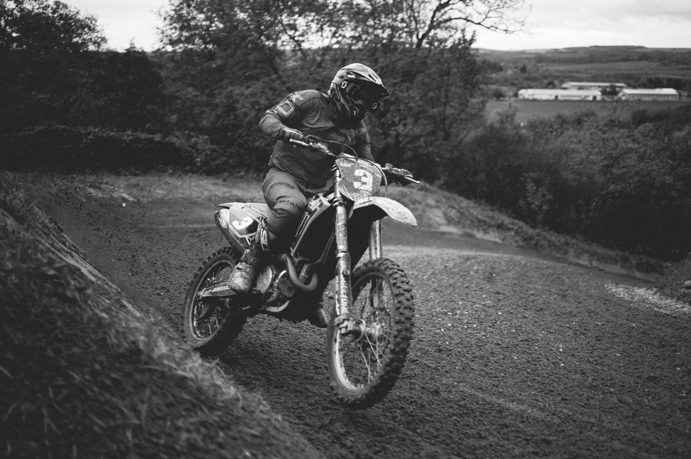 Black and white photo of a motocross rider in helmet and gear leaning a dirt bike through a muddy turn on a hillside track.