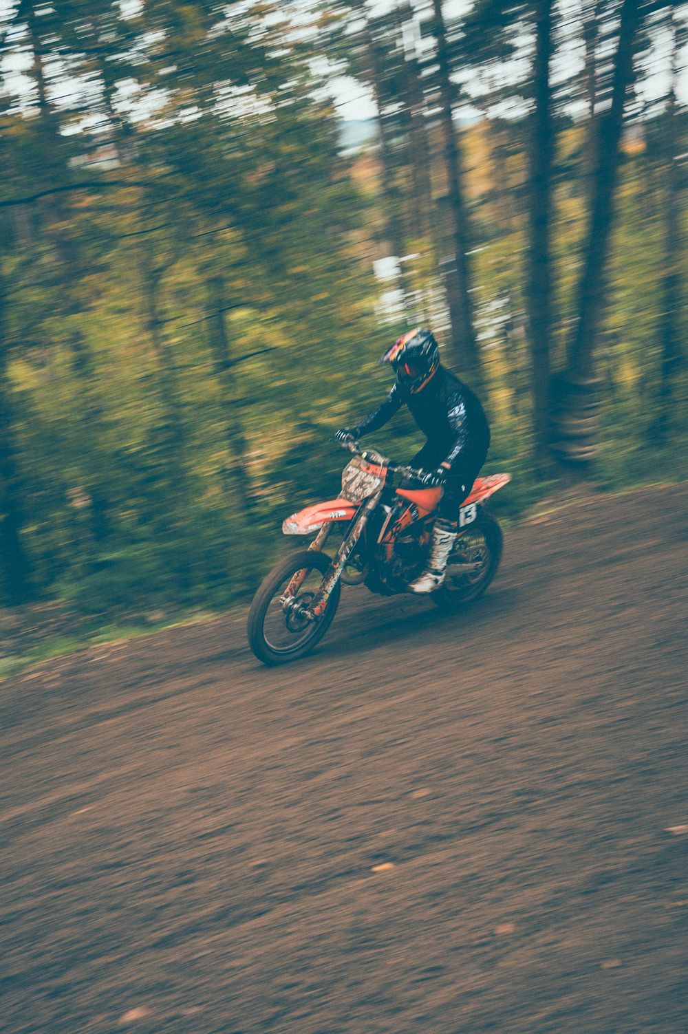 A motocross rider in a black helmet and gear speeds an orange dirt bike along a muddy forest trail with motion blur.