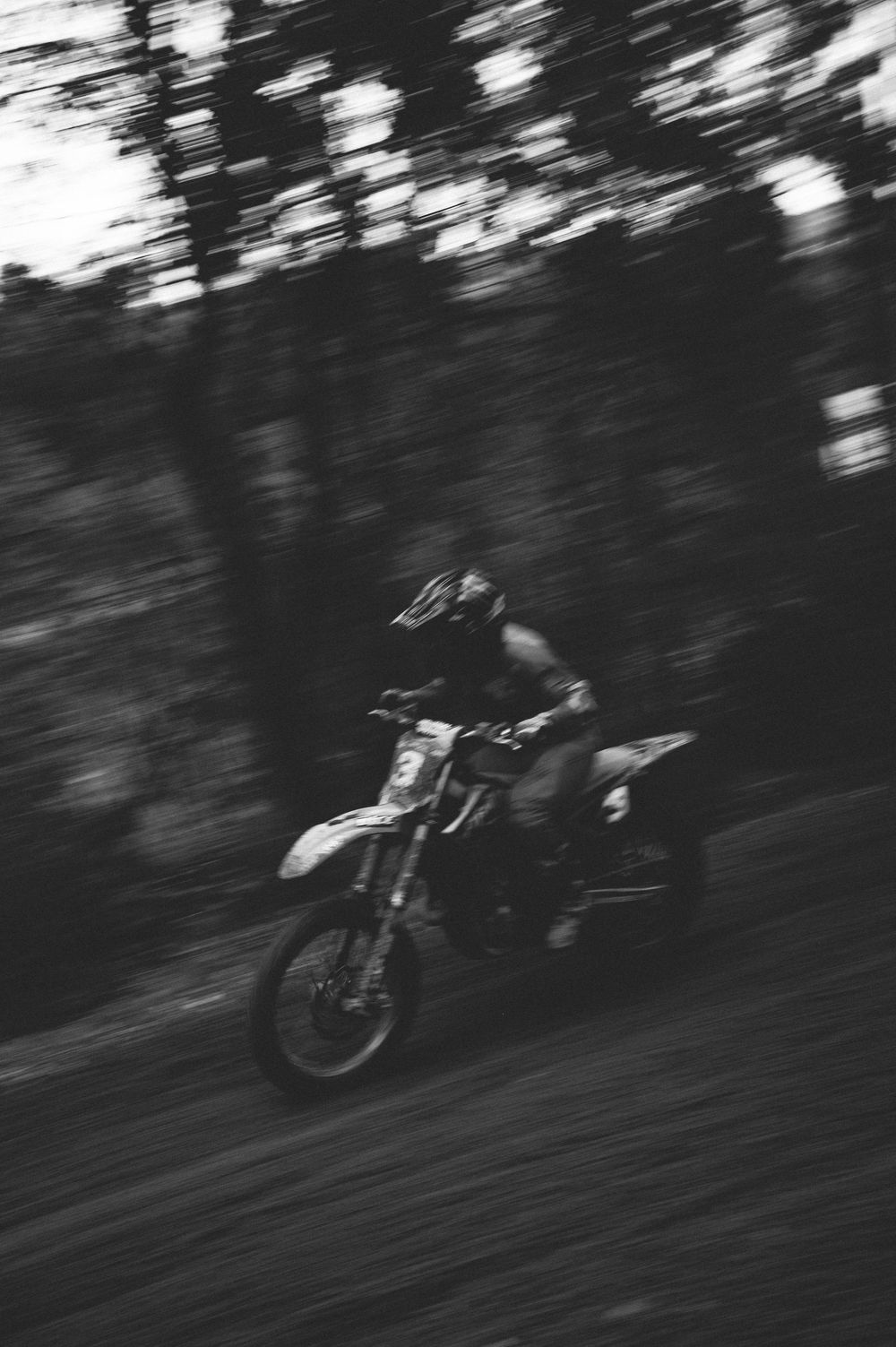 Black and white motion-blurred photo of a dirt bike rider in a helmet speeding along a forest trail.