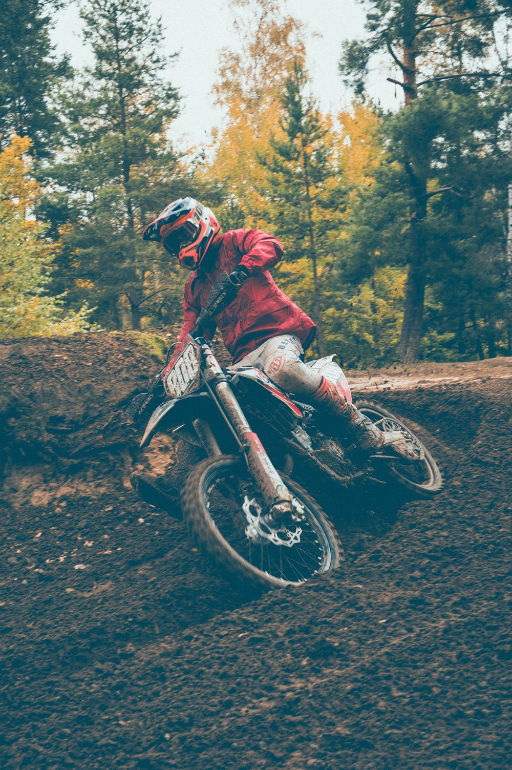 Motocross rider in a red jersey and helmet leans a dirt bike through a muddy forest track surrounded by autumn trees.