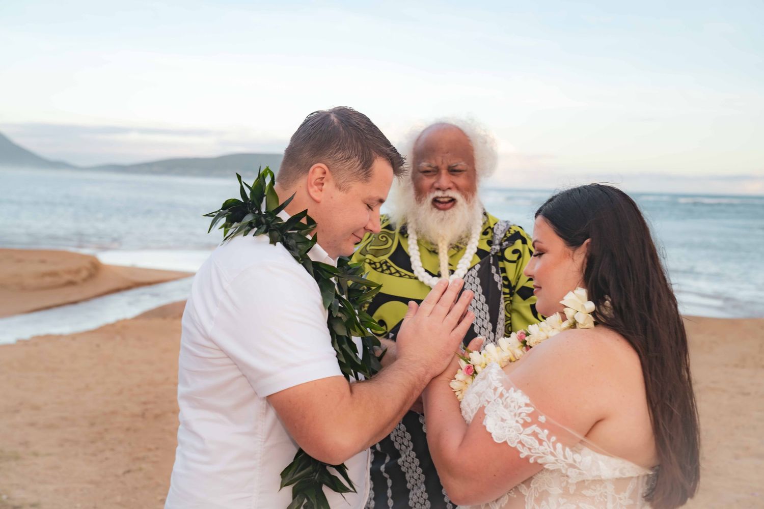 Couple exchanging vows on a beach during a Hawaiian wedding ceremony as an officiant in a green floral shirt and white beard blesses them.