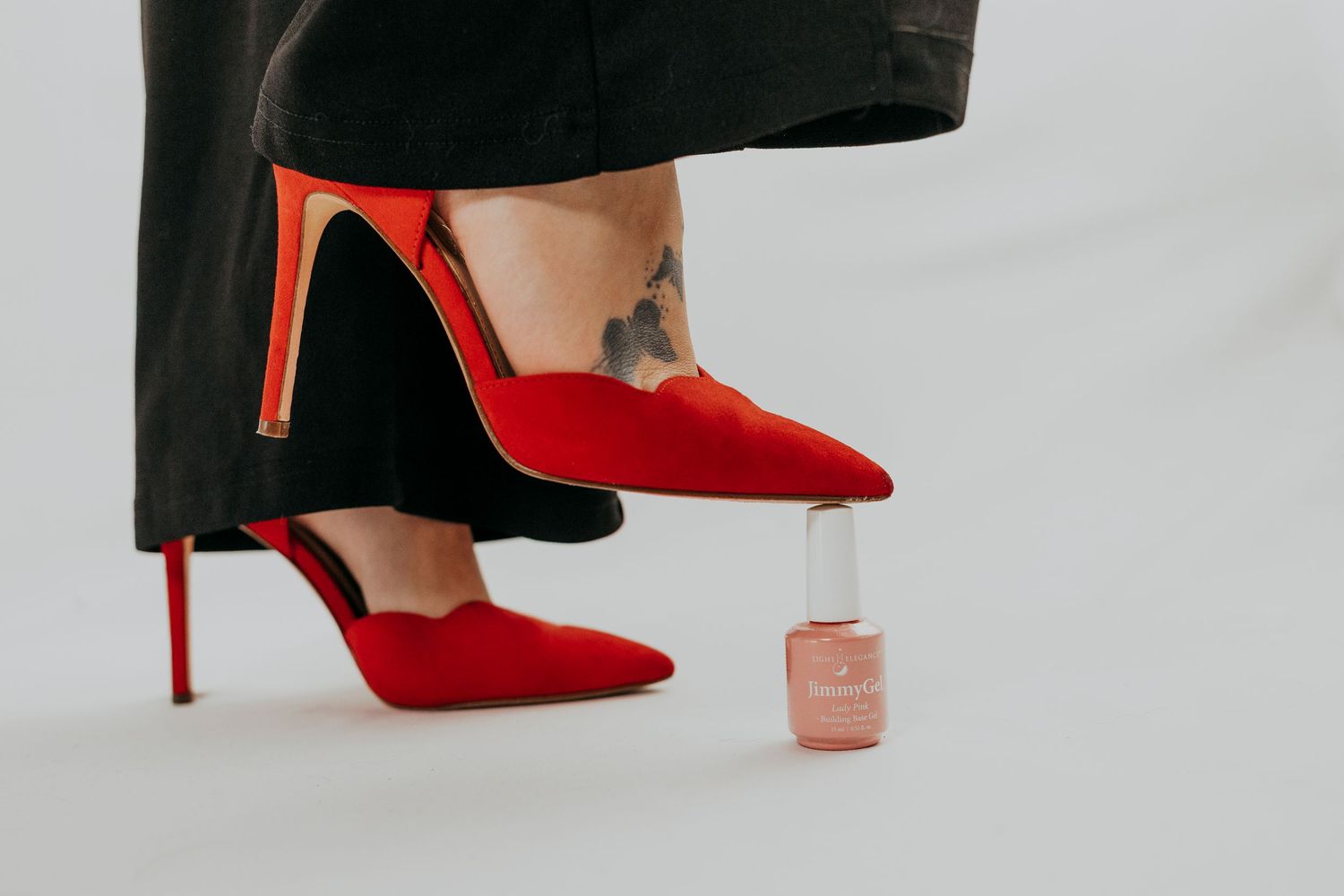 Woman in red pointed-toe stiletto heels with an ankle tattoo, standing beside a pink nail polish bottle on a light background.