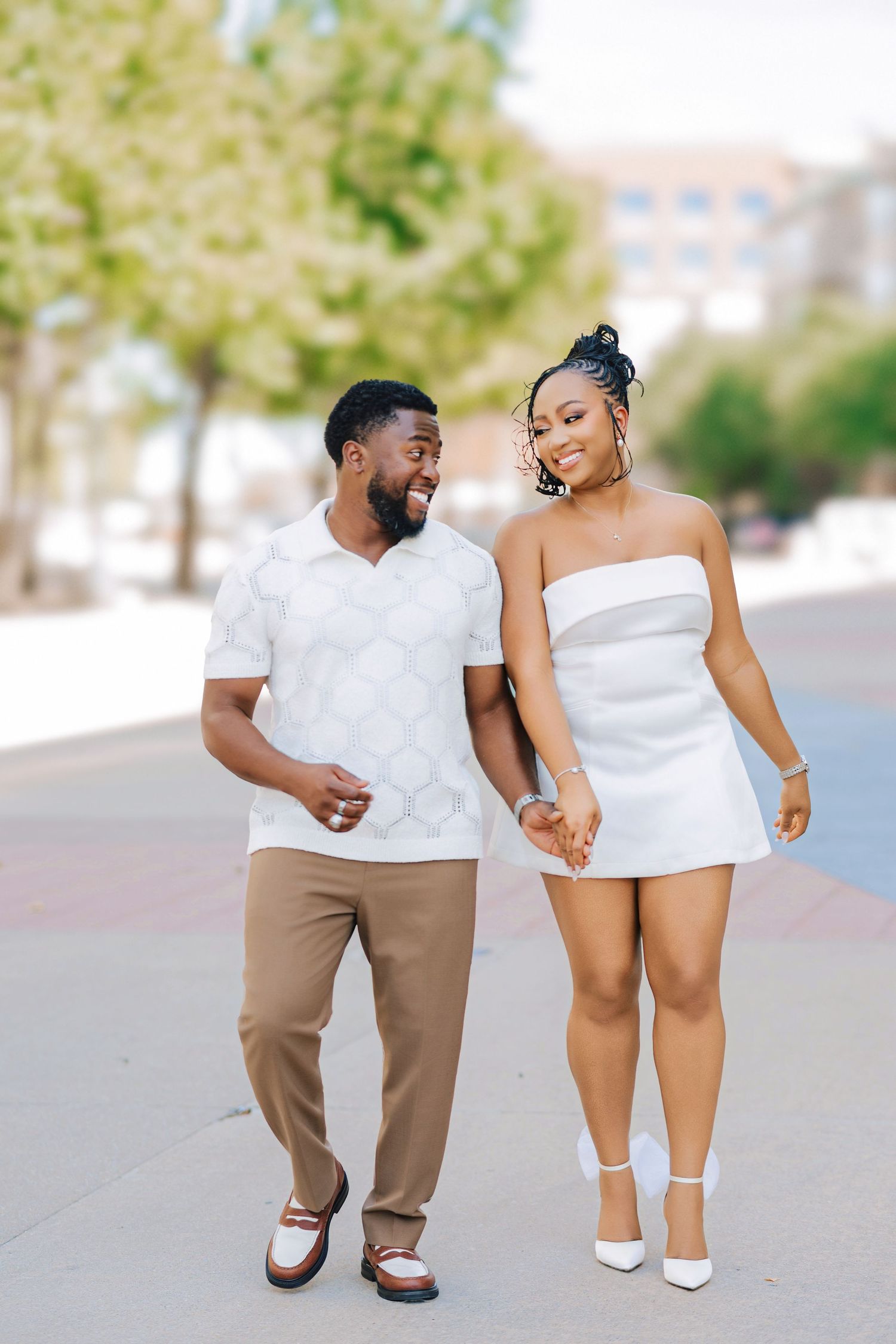 Smiling couple holding hands and walking down a sunny city sidewalk, he in a white polo and tan pants, she in a white strapless dress.