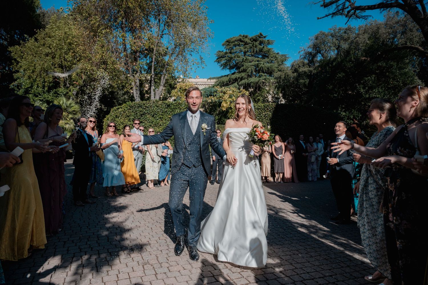 Smiling bride in a white gown and groom in a navy suit walk hand in hand past cheering wedding guests along a sunlit garden path.