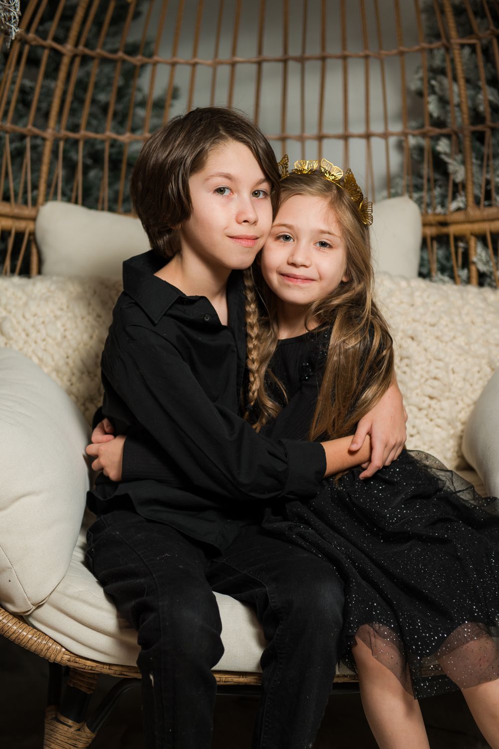 Two smiling children in black outfits hug on a cozy cream cushioned wicker chair during a festive holiday photo session.