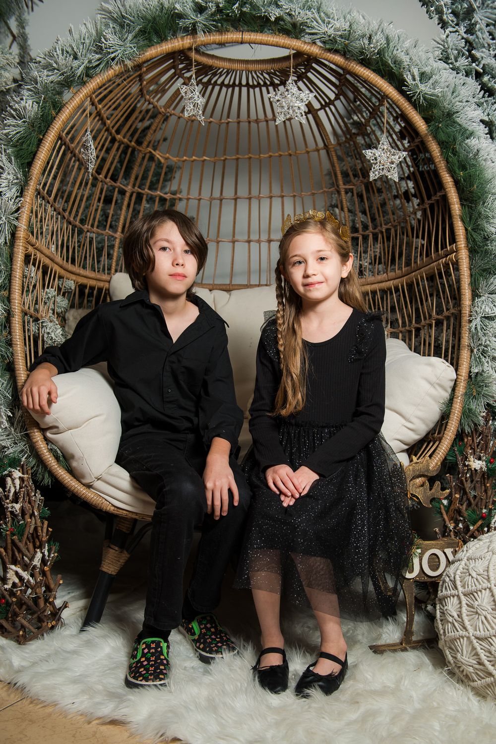 Boy and girl in black outfits sitting together in a rattan egg chair with a frosted holiday wreath and white faux fur rug.