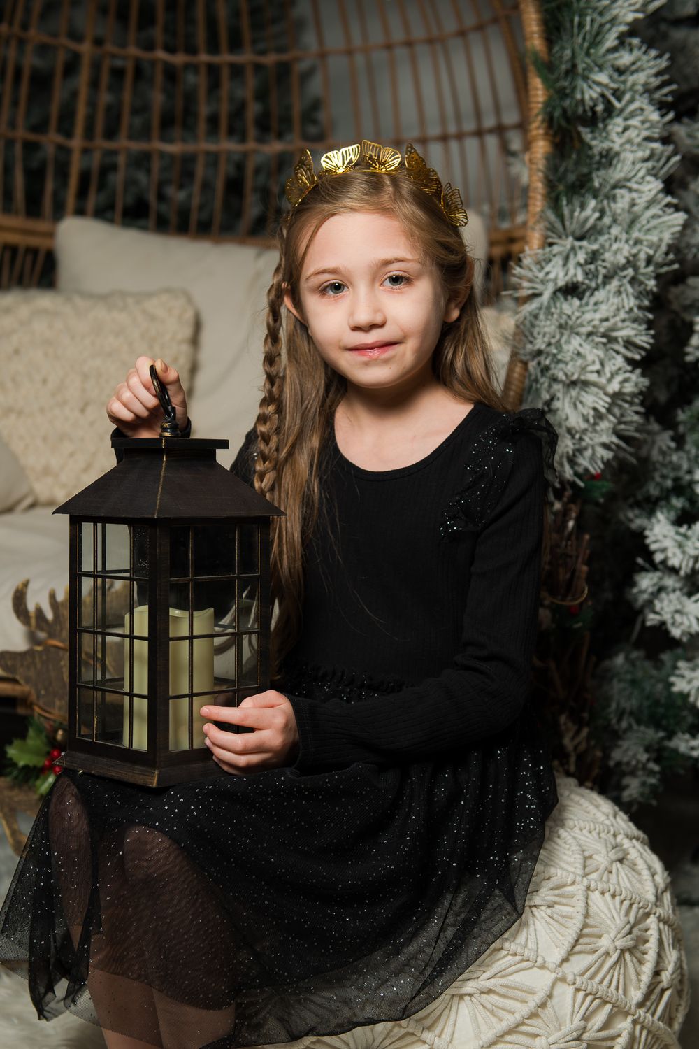 Young girl in a black sparkly dress and gold crown holding a black lantern with a candle beside a frosted holiday tree.