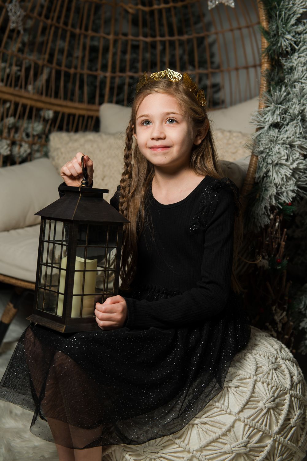 Smiling girl in a gold tiara and sparkly black dress holding a black lantern with candles beside a snowy holiday wreath.