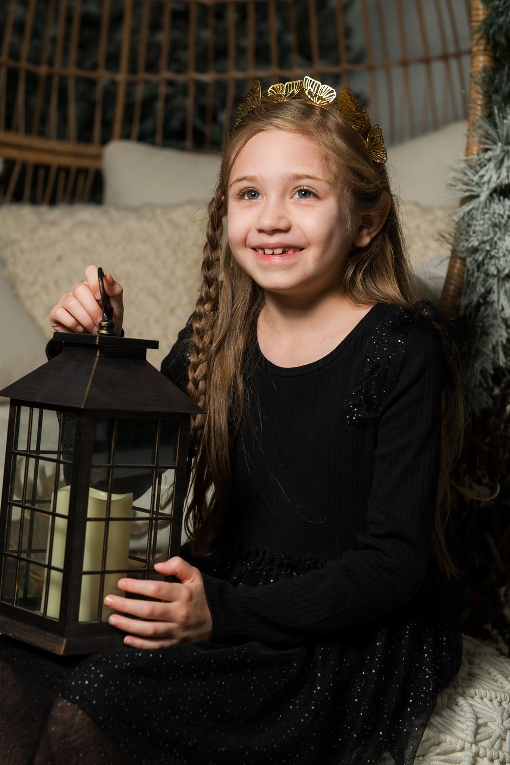 Smiling girl in a black dress and gold crown holds a lantern while sitting in a cozy winter holiday photo setup.