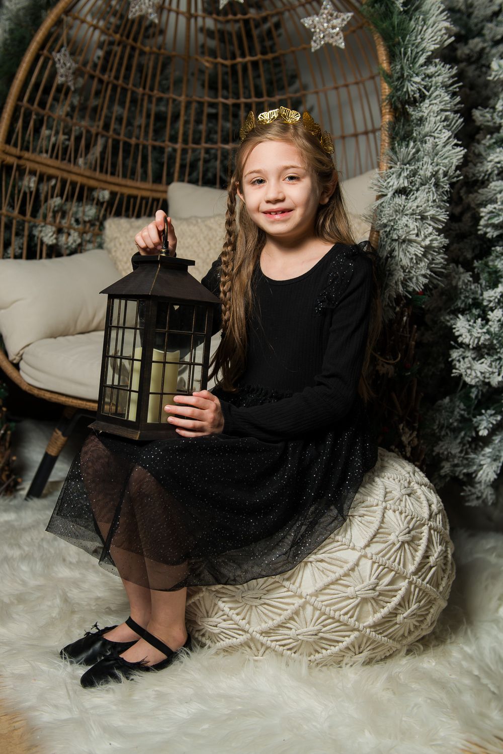 Girl in a black sparkly dress and gold crown sits on a knit pouf holding a lantern beside a frosted tree and rattan chair.