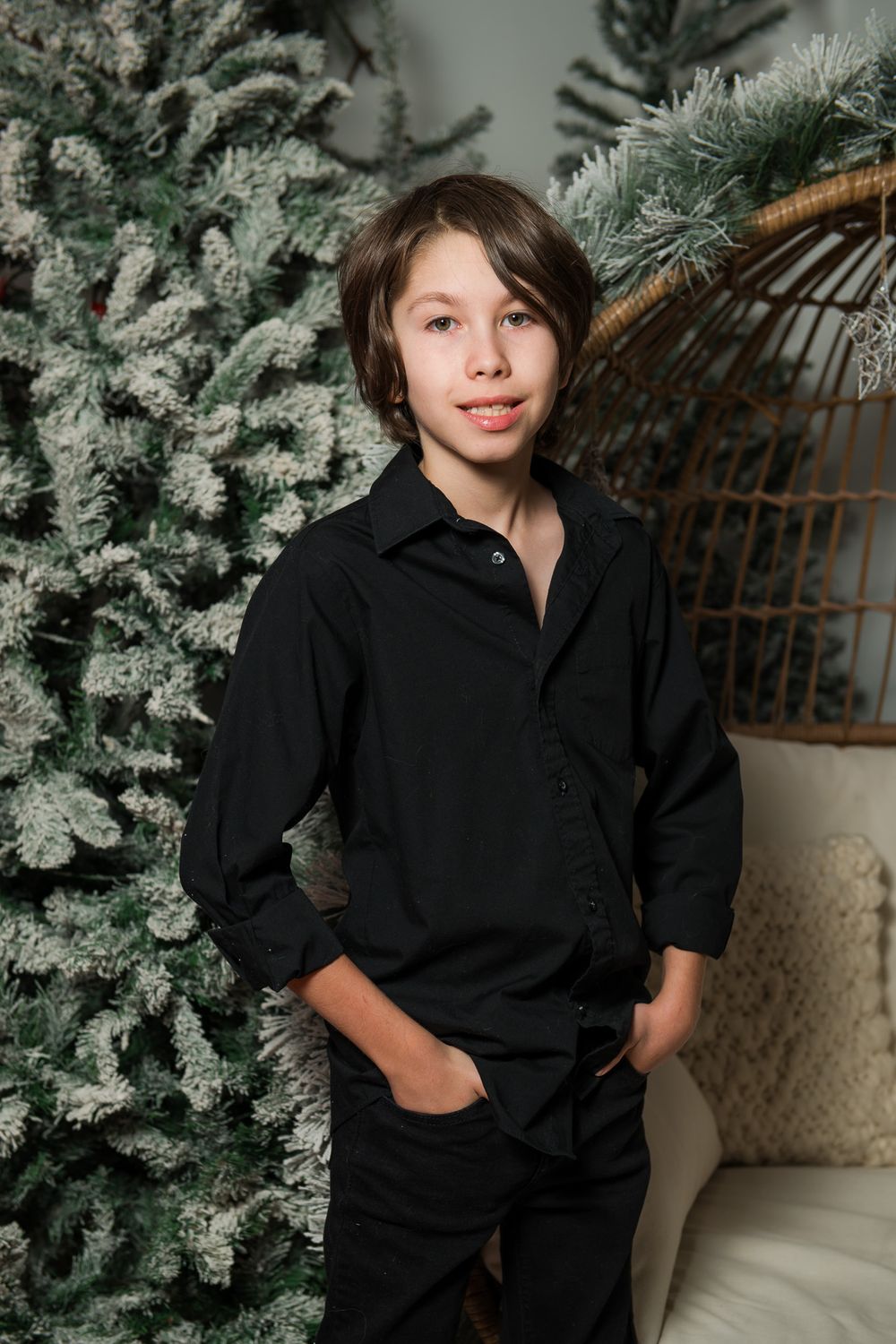 A boy in a black shirt and pants smiles with hands in his pockets in front of snowy flocked Christmas trees and a cozy cushioned chair.