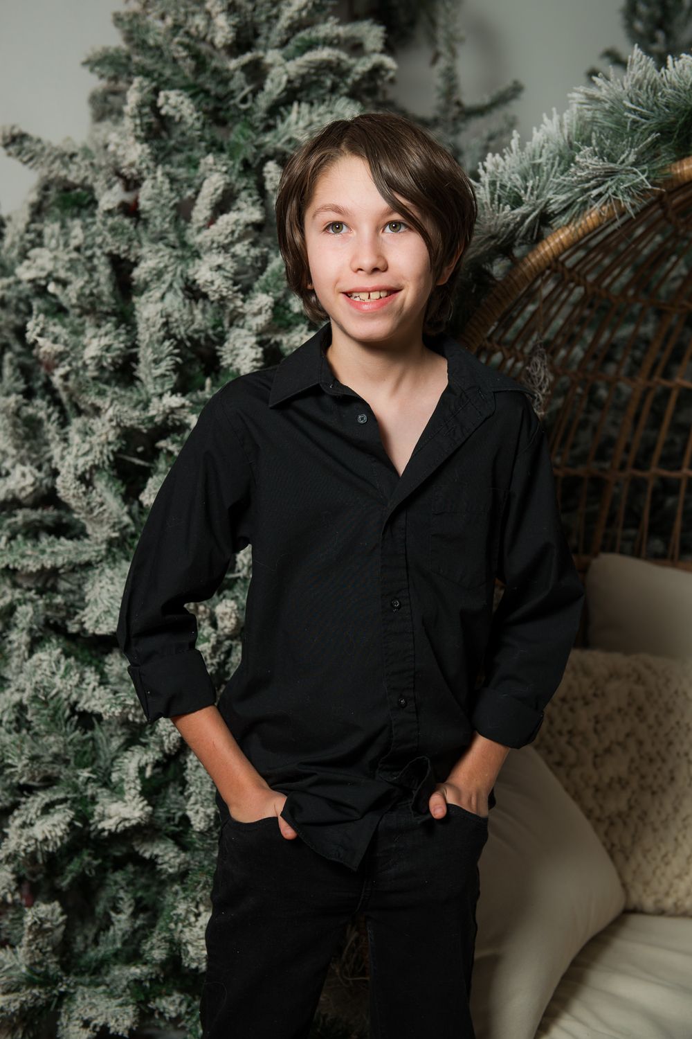 Smiling boy in a black button-up shirt and black pants stands with hands in pockets beside a flocked Christmas tree and a wicker chair.