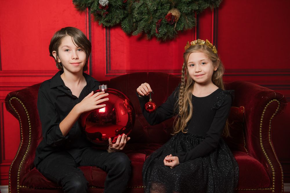 Boy and girl in black sit on a red velvet sofa holding a large red ornament under a Christmas wreath on a red wall.