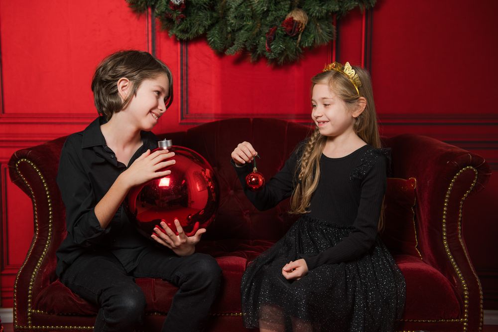 A boy and a girl in black sit on a red velvet sofa holding red ornaments in front of a red wall with a Christmas wreath.