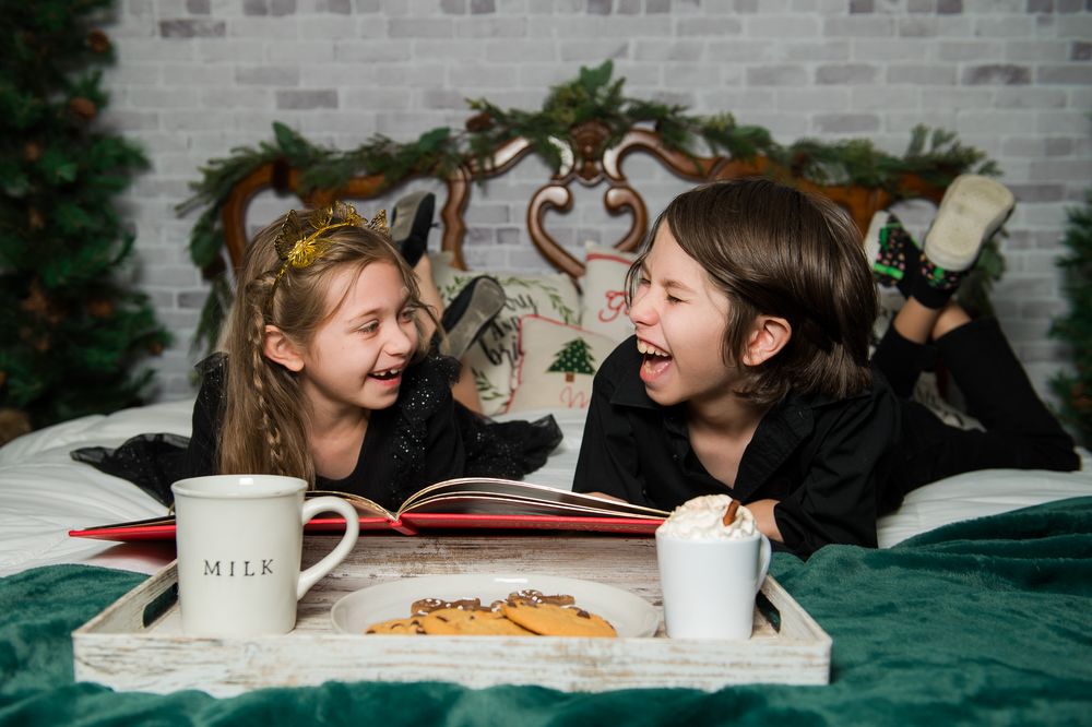 Two laughing children lying on a green bed reading a red storybook with milk mugs and cookies on a tray, Christmas trees behind.