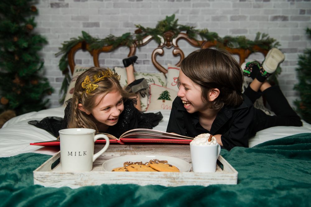 Two children in a golden crown and black clothes lie on a green bed with an open book, milk and cookies for a Christmas photo.