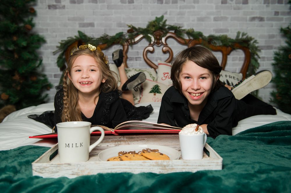 Two smiling children in black outfits lie on a green blanket reading a book with milk and cookies in a cozy Christmas photo setting.