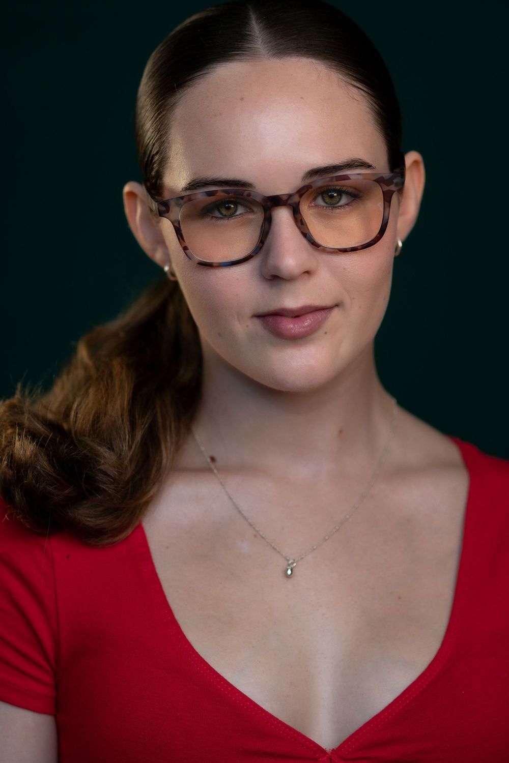 Professional portrait featuring a woman in a red v-neck top wearing tortoiseshell glasses against a dark backdrop.