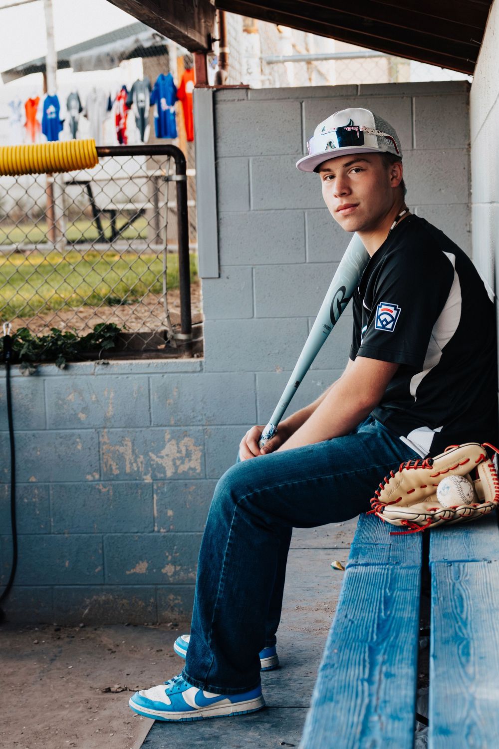 A baseball player sits in the dugout wearing a cap and uniform while holding food during a game break.