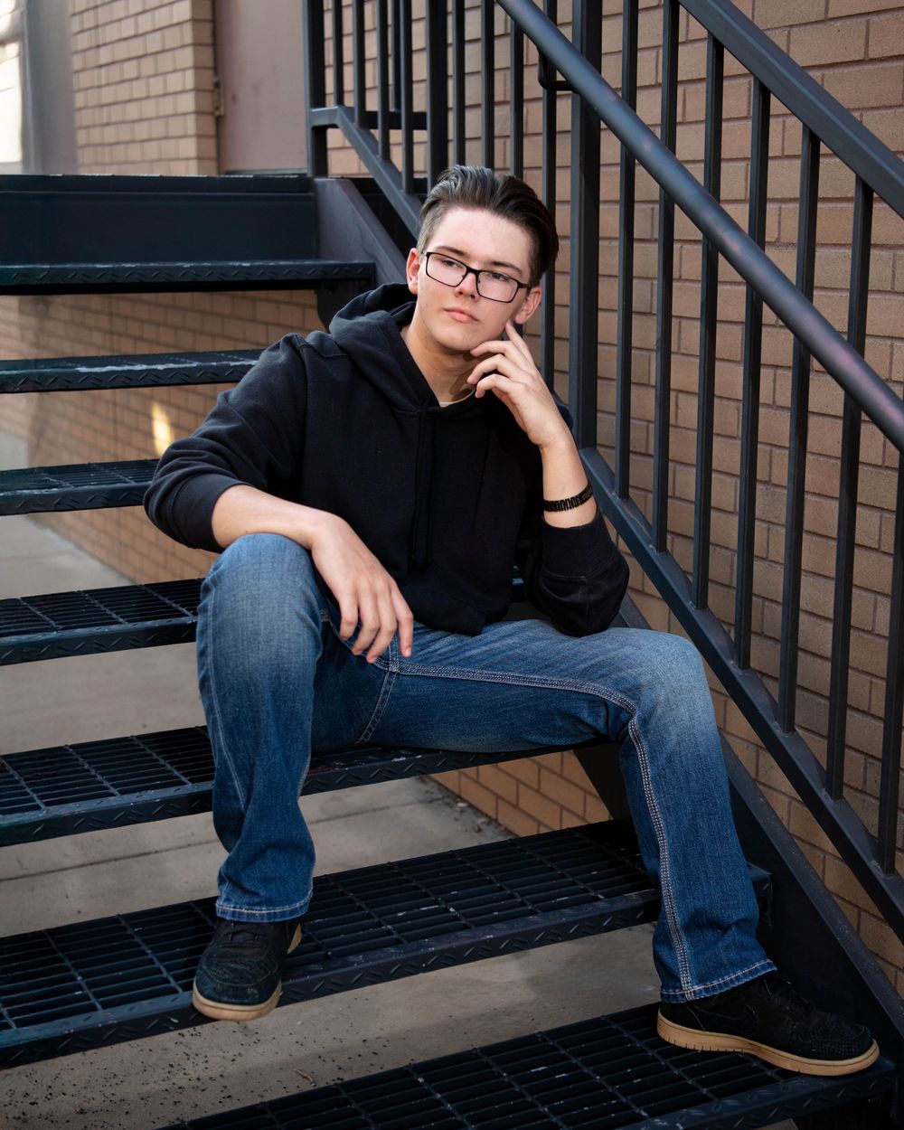 Person in casual attire sitting on concrete steps against a brick wall wearing glasses and dark clothing.