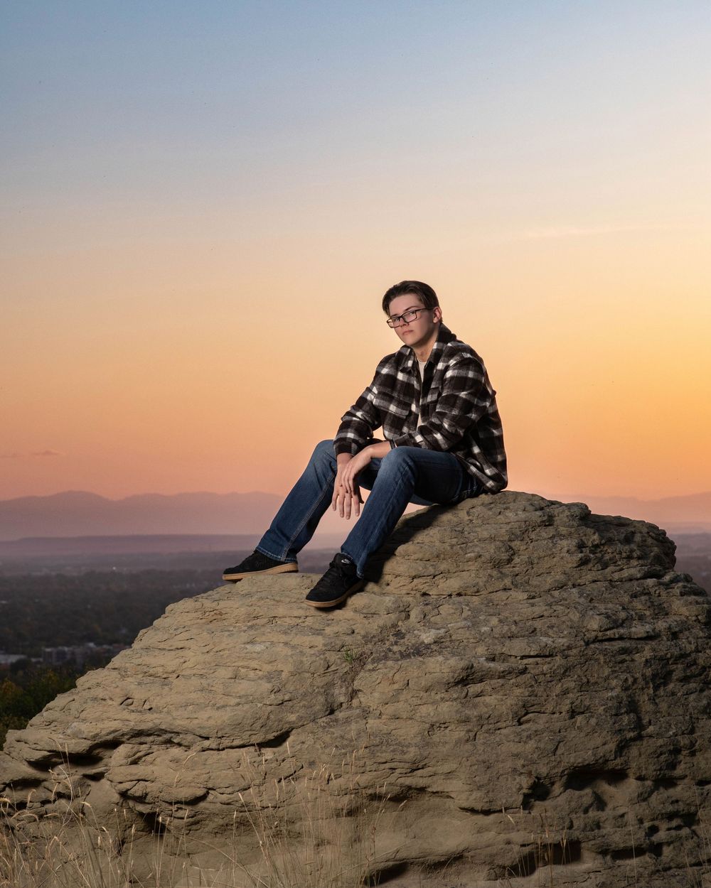 A person sits on a rocky outcrop overlooking a scenic mountain landscape at sunset.