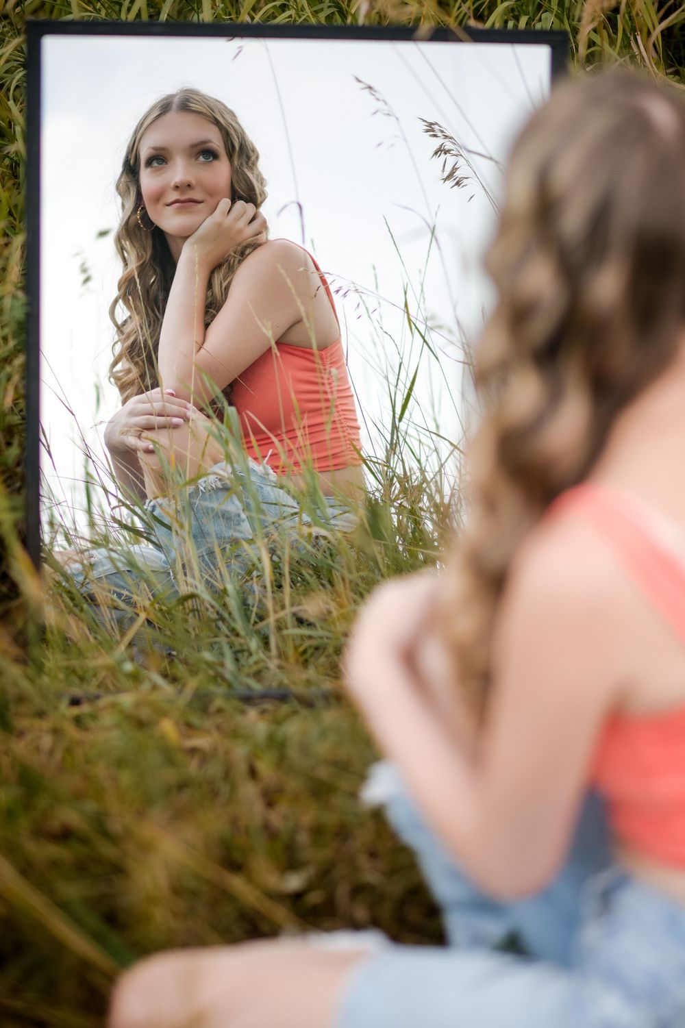 A person in a coral top and jeans sits in tall grass during a sunny outdoor photo session.