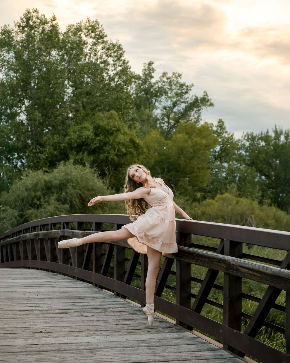A dancer performs graceful poses on a wooden bridge during sunset with trees in the background.