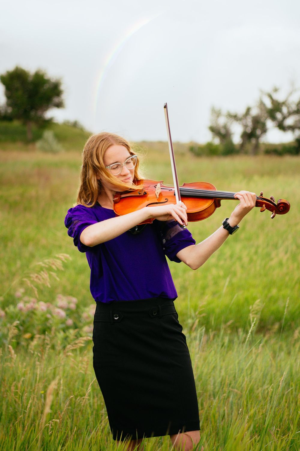 Musician in a purple top and black skirt plays violin outdoors in a grassy meadow during golden hour.