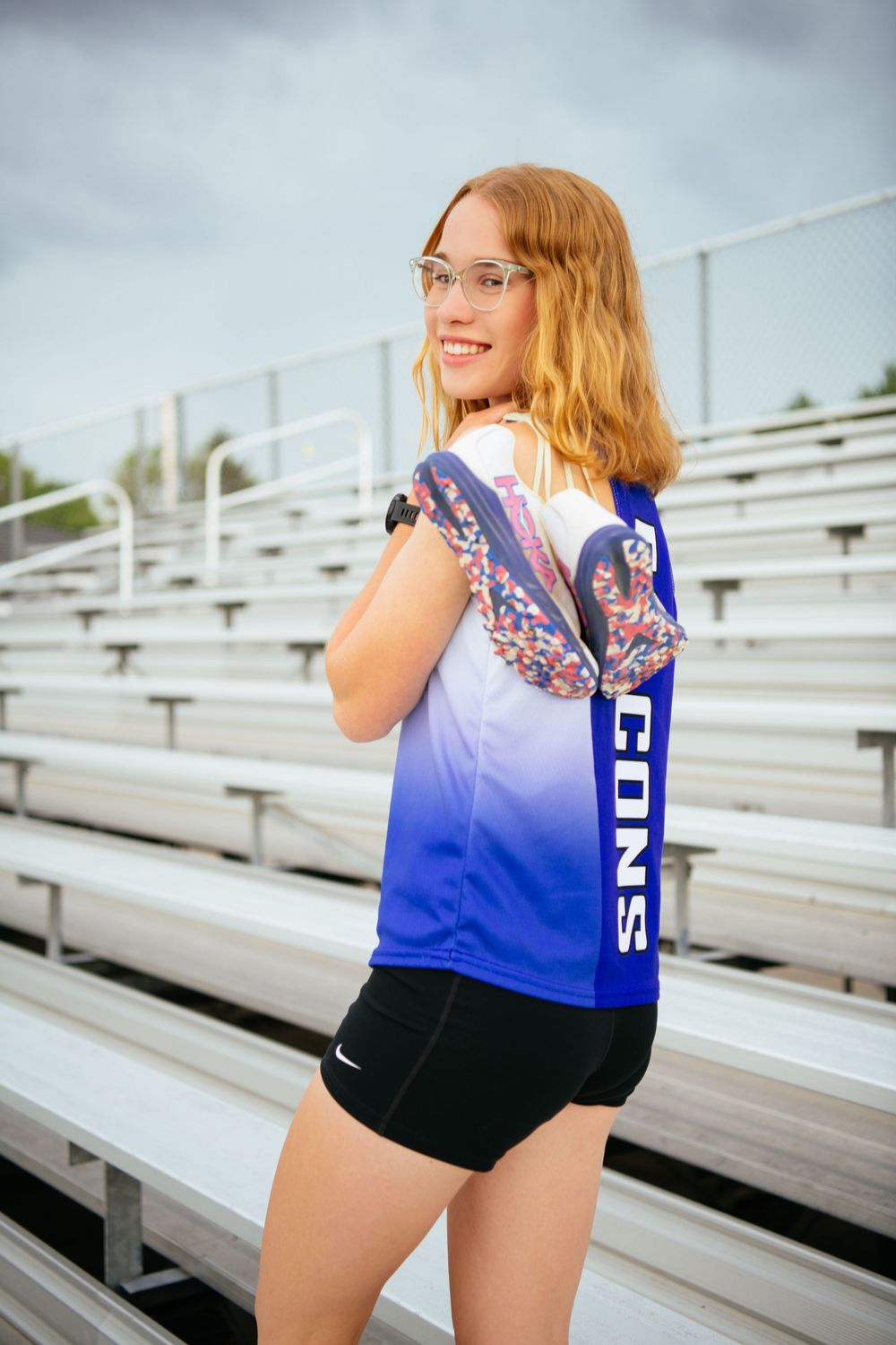 A person in a blue and white top and black shorts poses playfully on bleacher seats at an outdoor stadium.