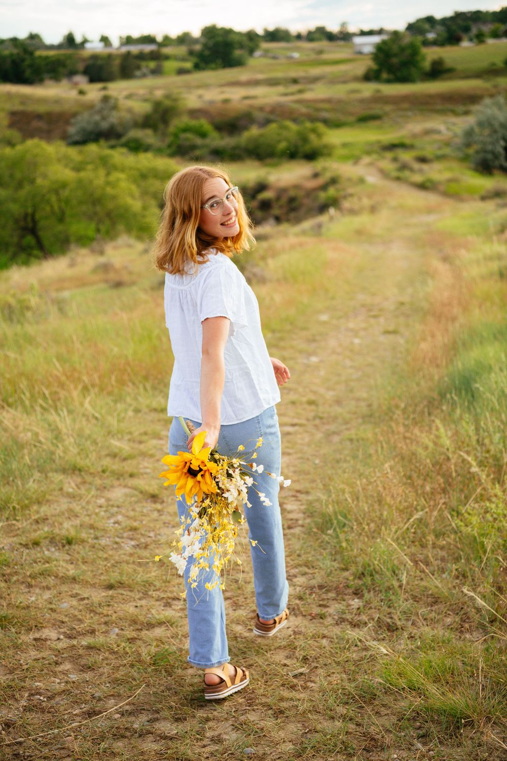 A person in jeans and white shirt walks along a grassy path while holding bright yellow sunflowers on a sunny day.