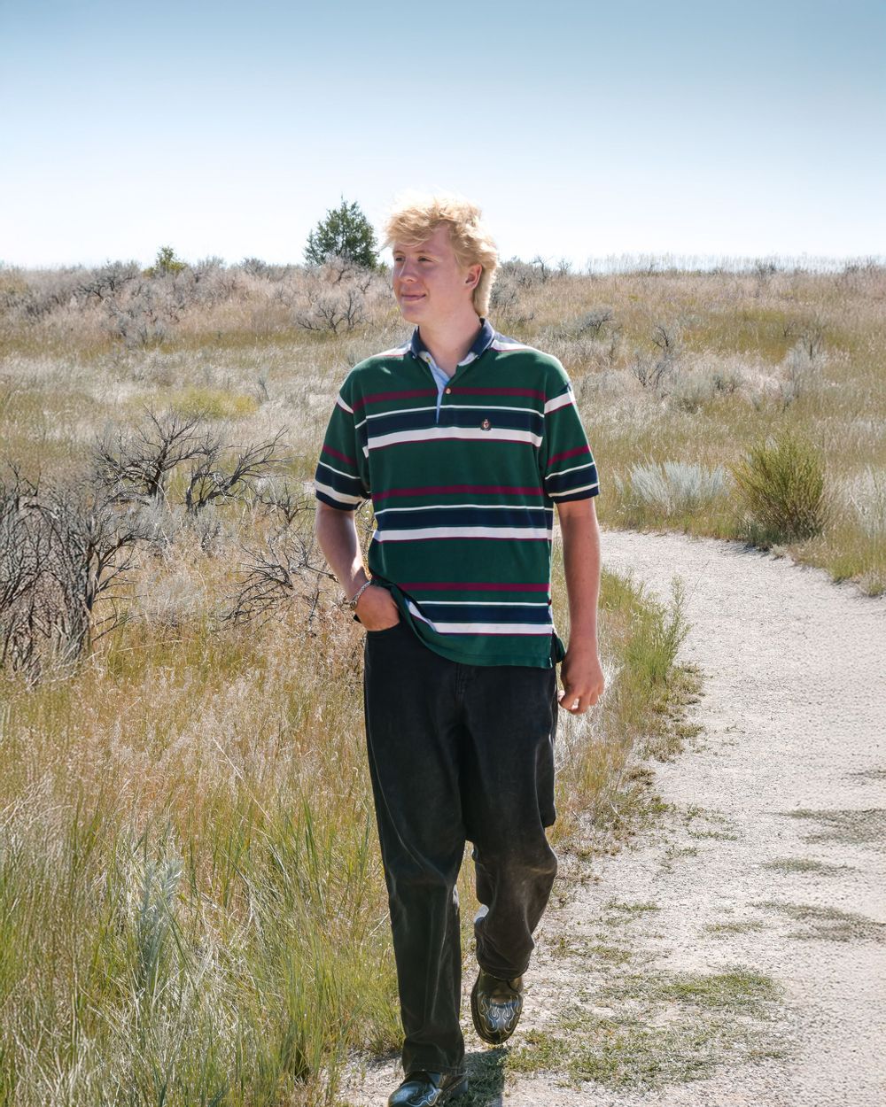 Someone wearing a green striped polo shirt walks along a winding dirt path through dry grassland terrain.