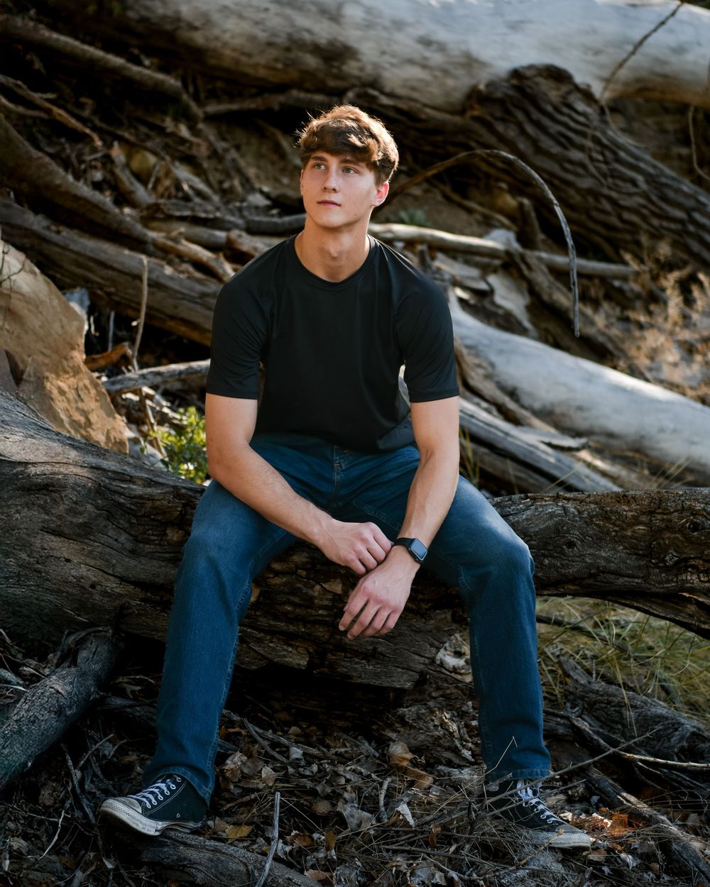 A person in casual attire sits on a fallen log in a wooded outdoor setting during golden hour lighting.