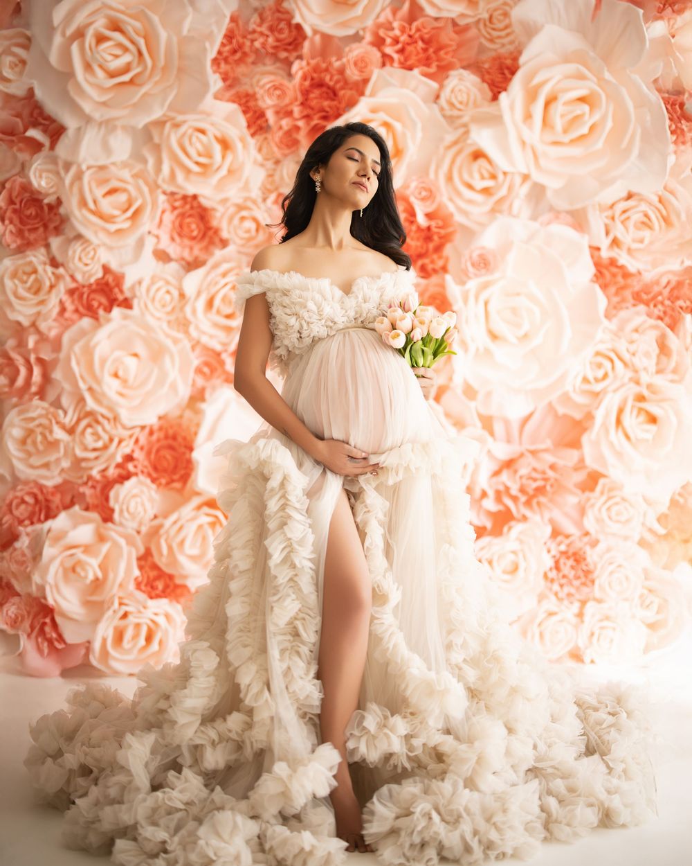Elegant maternity portrait in front of a romantic coral and peach rose flower wall backdrop featuring a flowing white gown.