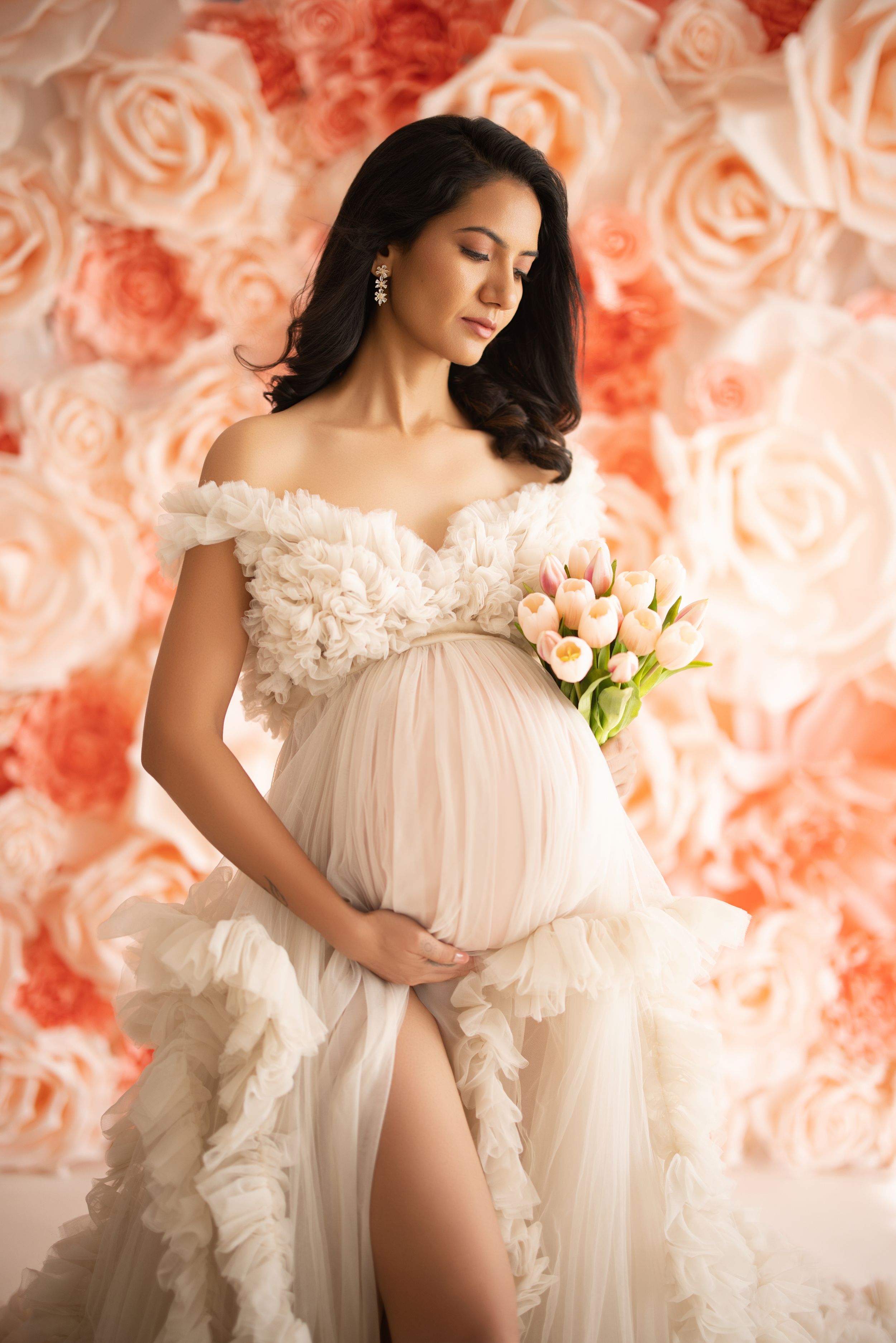 Maternity photoshoot featuring flowy white sheer gown with ruffles against coral-toned background.