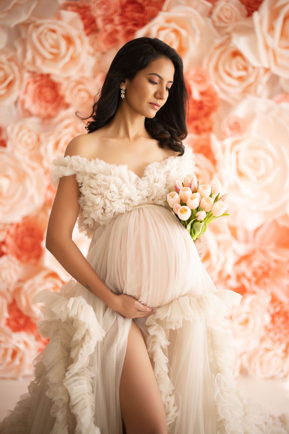 Maternity photoshoot featuring flowy white sheer gown with ruffles against coral-toned background.