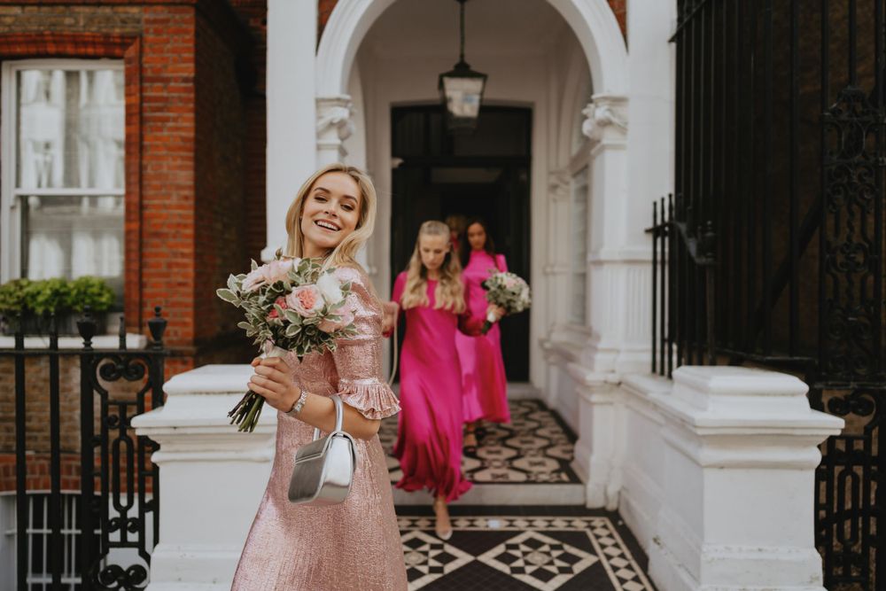 Person in pink dress holding flowers outside elegant brick building entrance.