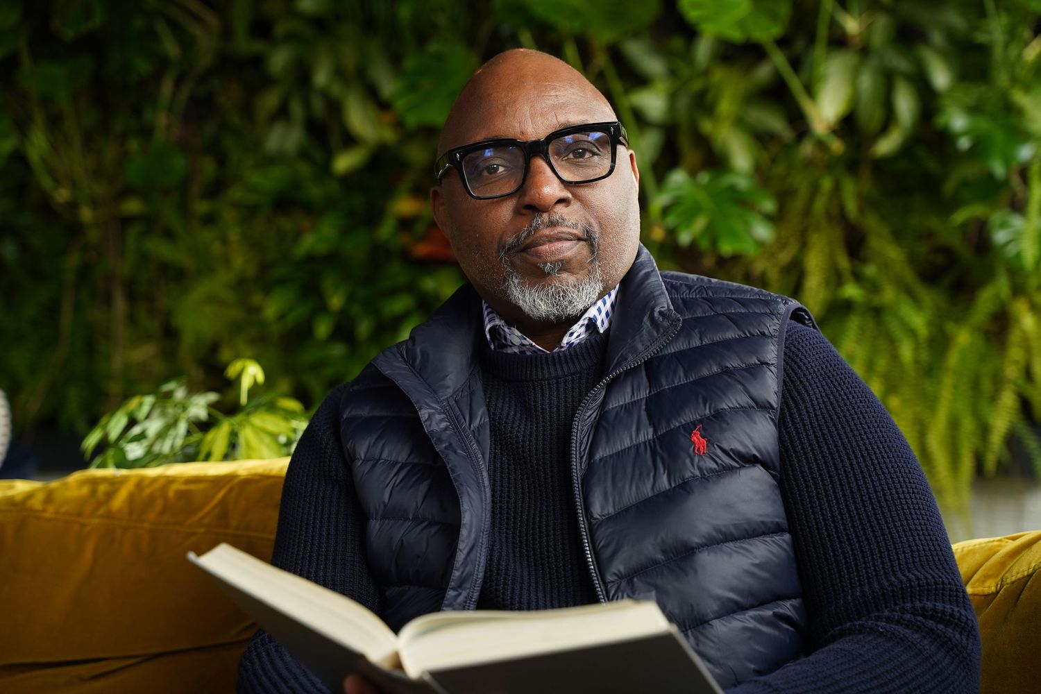 A black person in glasses and a puffy vest sits comfortably on a yellow bench while reading a book outdoors.