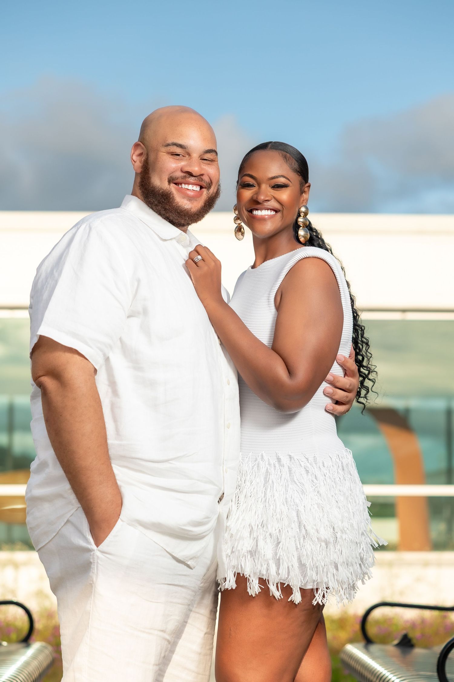 A couple wearing white outfits share romantic moments on an outdoor balcony with an ocean view in the background.