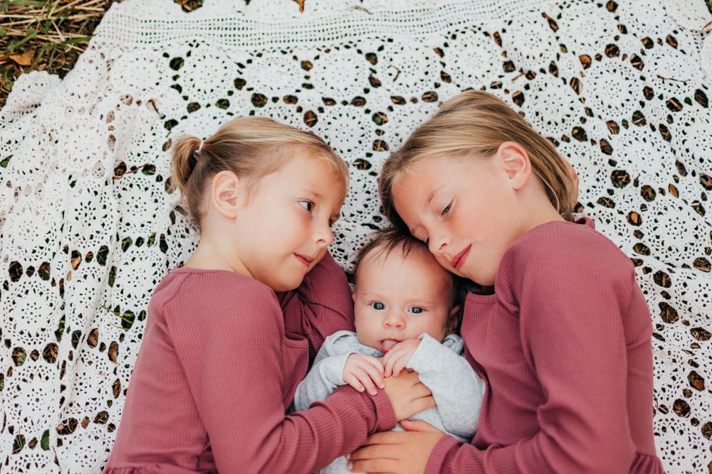 Three siblings wearing pink and light blue tops laying together on a white lace crochet blanket.