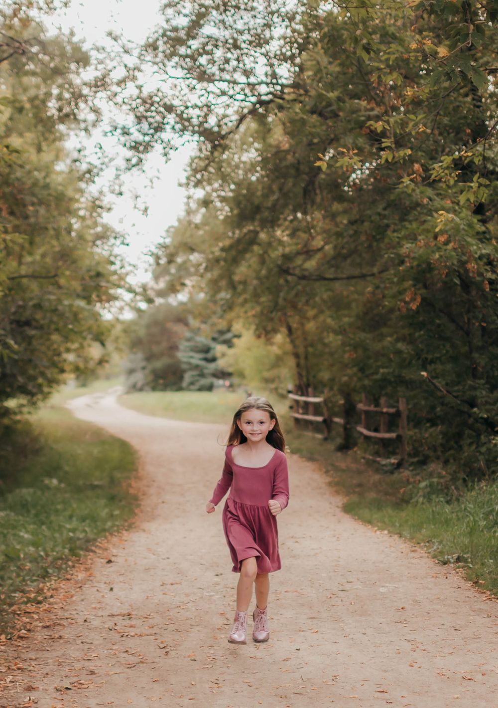 A small child wearing a burgundy dress walks along a winding dirt path surrounded by autumn trees.