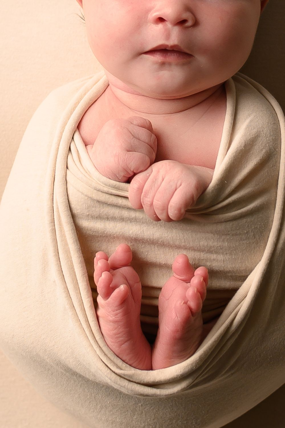 Tiny infant hands and toes peek out from a cozy beige swaddle wrap in a gentle newborn photo series.