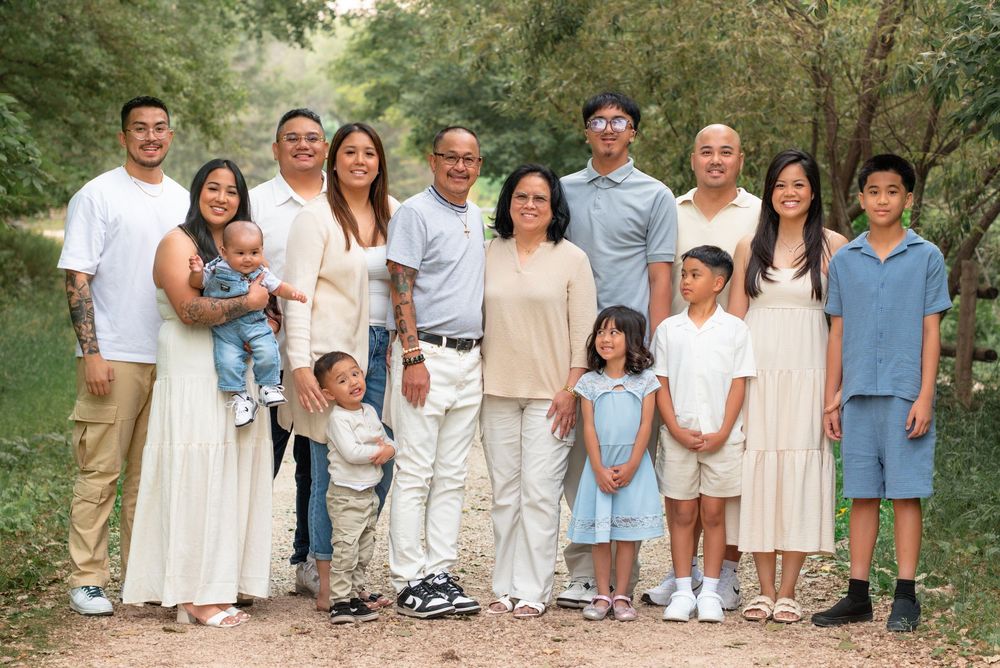 Large family group in coordinated white, beige and blue outfits poses together on outdoor path surrounded by trees.