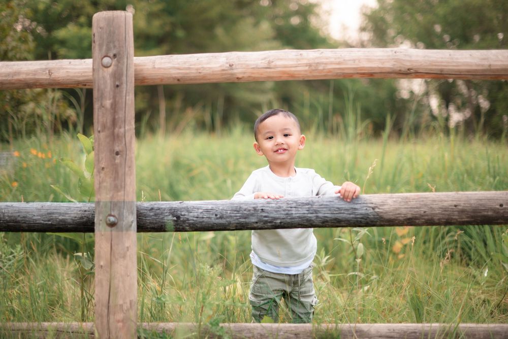 Young child in white shirt smiling while standing at rustic wooden fence in green meadow at sunset.