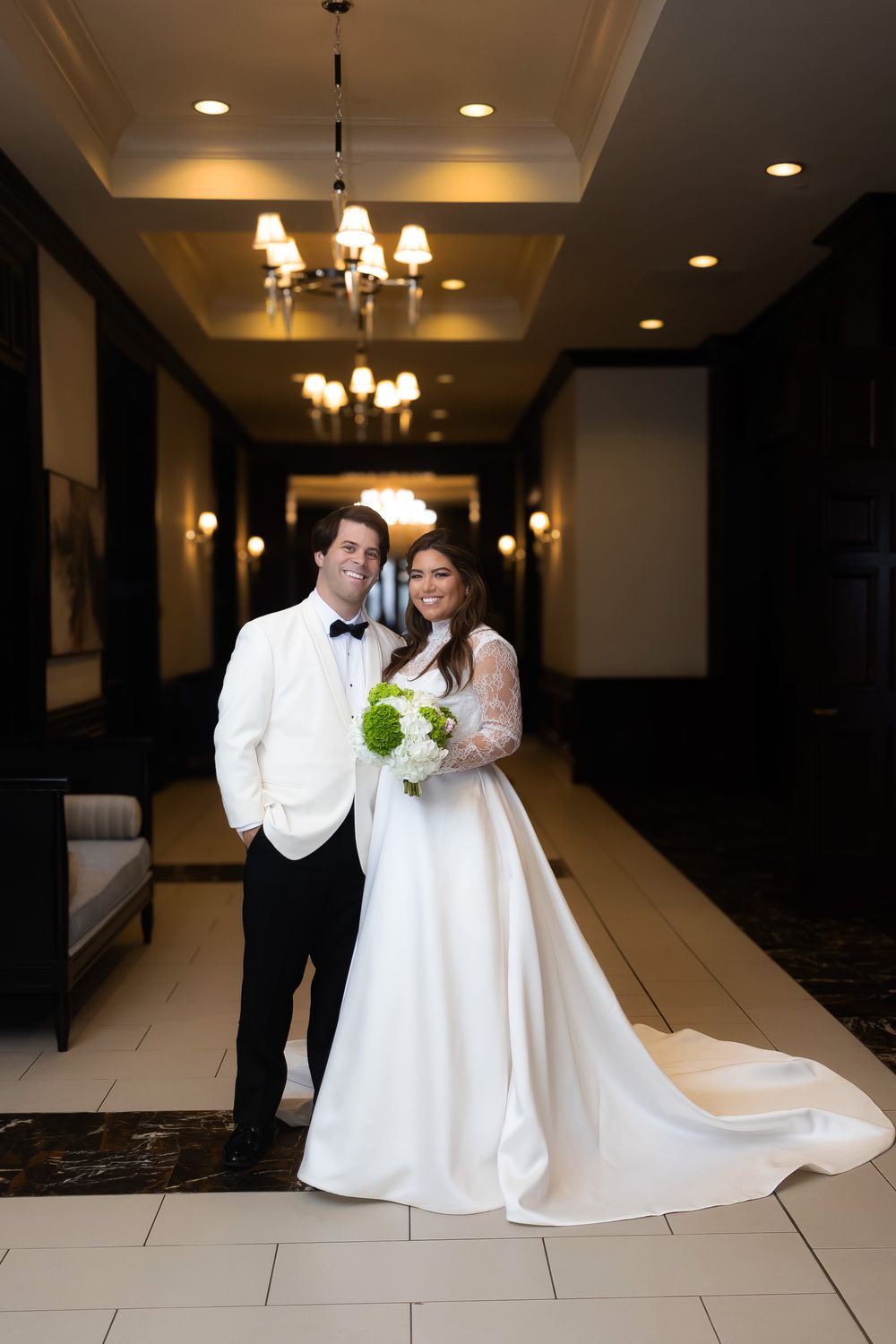 A wedding couple poses in an elegant hotel hallway with chandeliers and a long white train on the bride's gown.