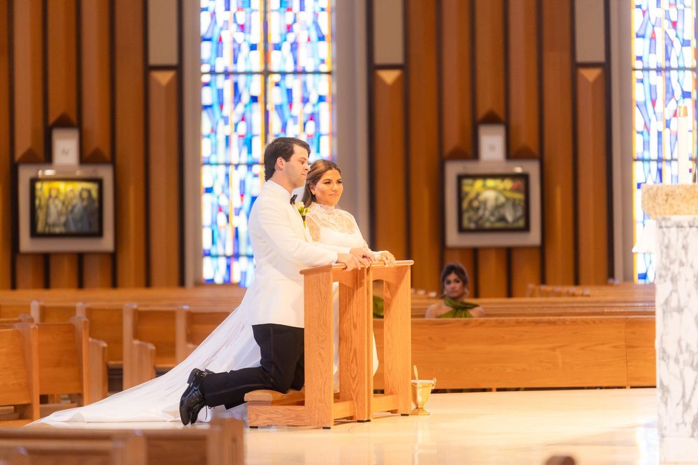 A couple in wedding attire kneels at a wooden altar in a church with stained glass windows and wooden paneling.