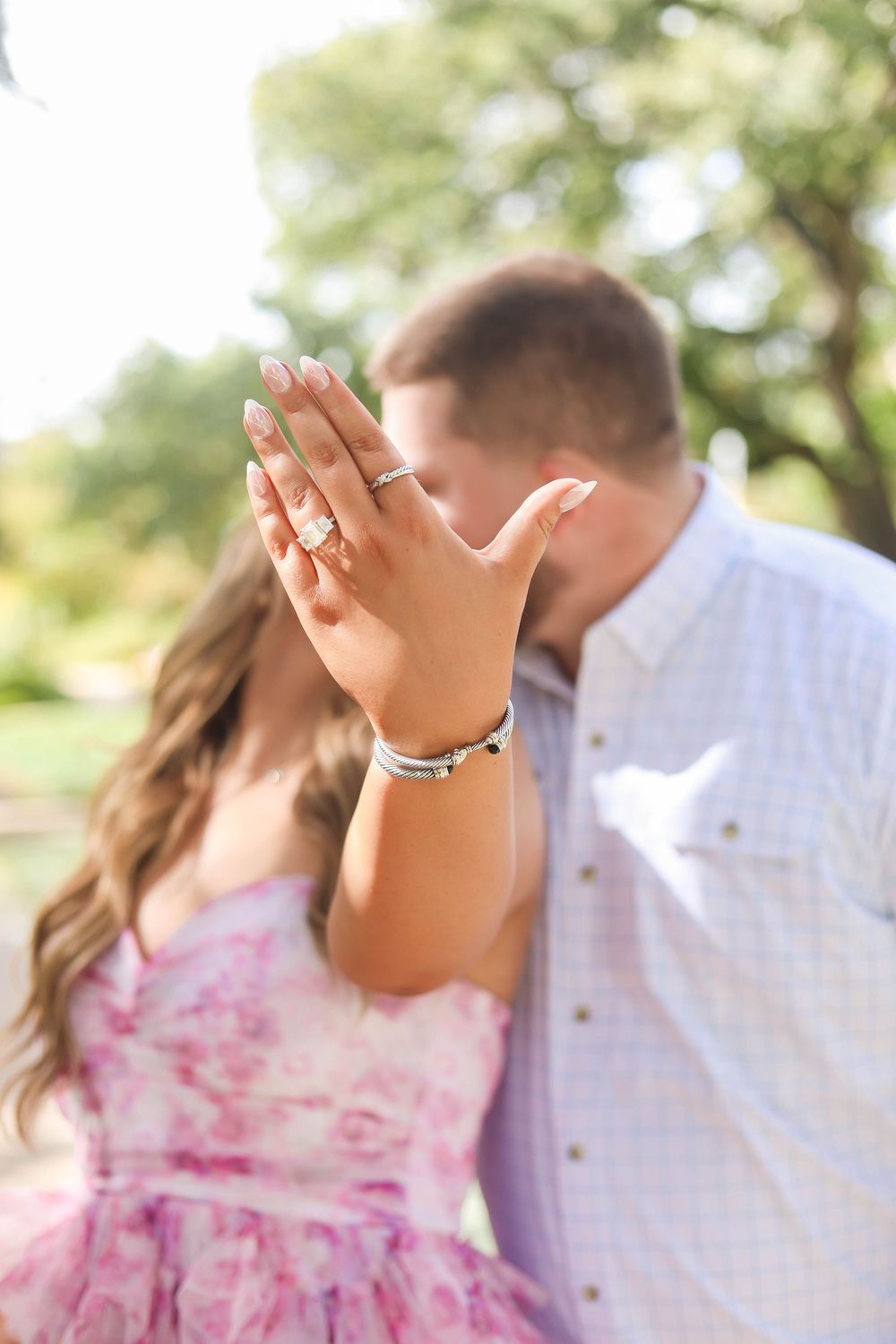 A couple in love shares a romantic moment outdoors, with her in a pink floral dress and him in a white shirt.