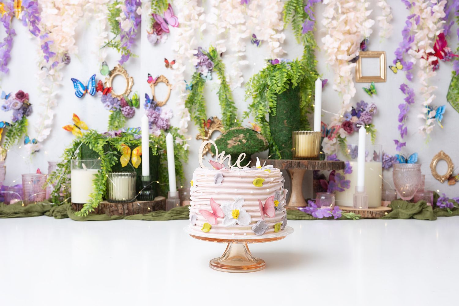 Pink floral cake on stand with butterfly decorations against whimsical garden backdrop.
