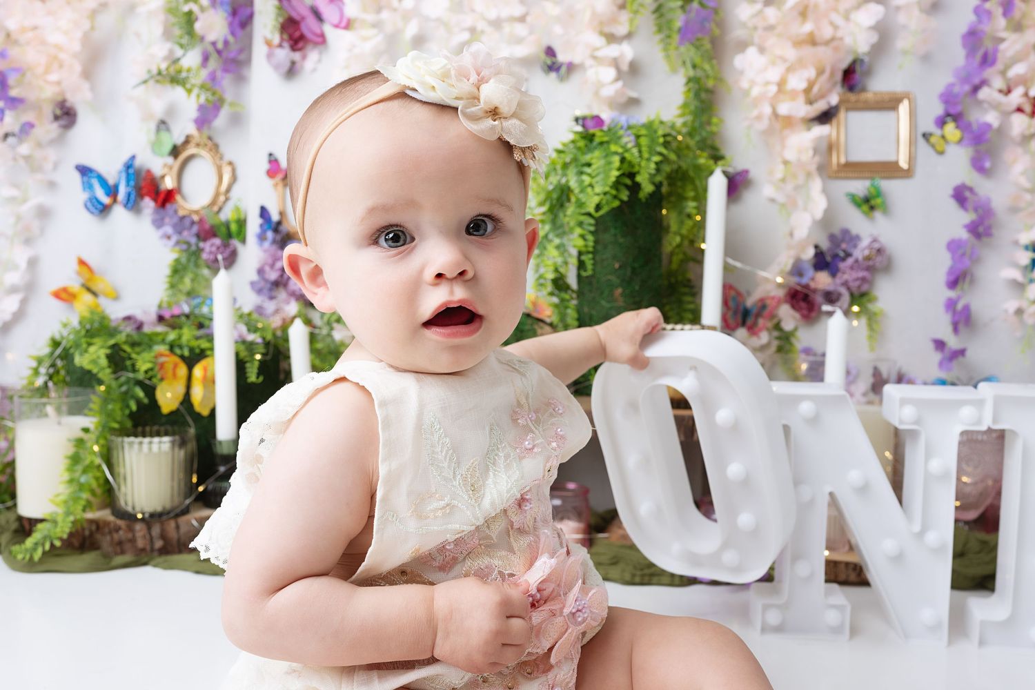 Baby in cream dress poses with decorative letters against floral backdrop.